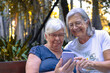 © luciano - Two senior women smiling and enjoying a sunny day outdoors, sharing a digital moment and connecting with a smartphone, representing an active retirement