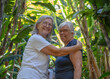 © luciano - Two cheerful senior adult women embracing in a vibrant tropical garden, sharing a warm moment of genuine friendship and happiness while surrounded by lush green foliage