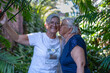© luciano - Two senior women embracing outdoors, one kissing the other on the cheek, sharing a moment of joyful friendship and affection in a lush garden setting
