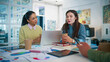 © ihorvsn - Female colleagues sitting at desk with laptop and papers. Team members discussing project details. Woman holding coffee cup and explaining ideas. Business people working together in glass office room.