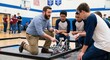 © whitestorm - Teacher guiding students in a robotics competition in a school gymnasium, focusing on teamwork and STEM.