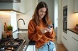 © Viktor - Smiling girl stands in modern kitchen typing message on phone. Young woman uses smartphone for communication or social media, browsing online. Casual home lifestyle.