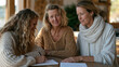 © forenna - Middle-aged therapist guides an adult daughter through a structured journaling exercise at a wooden table, generational patterns worksheet visible nearby, intergenerational trauma