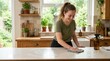 © Xenia - Smiling young woman wiping a white kitchen counter with a reusable cloth. Eco friendly household cleaning and zero waste lifestyle concept. Amber glass spray bottle and wooden cabinets in background