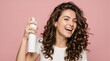 © ViskBx - Happy woman with curly hair holding a hairspray bottle, isolated on a pink background