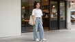 © Elijah - Full body portrait of stylish young Asian woman wearing white t-shirt and wide leg blue jeans carrying beige canvas tote bag standing outside modern cafe storefront showcasing