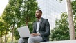 © AZ Studio - Young Black man in blazer working on laptop outdoors near modern building