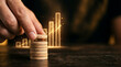 © IHK Creations S - A hand placing a wooden coin on top of a stack with glowing yellow bar graphs in the background representing financial growth and success