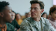 © CYBERPINK - Man with short, curly hair and glasses sitting in front of a group of people. he is wearing a light green jacket and appears to be deep in thought.