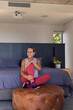 © wavebreak3 - Woman sitting on tufted ottoman in bedroom holding knee wearing lilac tank red leggings, projector