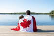 © DEXTEROUA - Father and daughter wrapped in large Canadian flag sitting on wooden dock by lake. Canada Day celebration outdoor. Dad and child sitting together on 1 of July celebrating national Canada Day.
