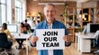 © MQ-Illustrations - Smiling senior caucasian man holding a Join Our Team sign in a modern diverse office