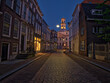 © Sami - Dordrecht, South Holland, Holland A lively European square at dusk, featuring historic buildings with glowing windows, street lamps, and outdoor cafes. People sit on benches.