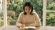 © AoIro scIA - 明るい部屋のテーブルでノートに日記や文字を書く若い日本人女性。おうち時間での勉強や丁寧な暮らしのライフスタイル写真