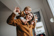 © qunica.com - A mother carries her toddler on her shoulders indoors, both wearing warm jackets. The playful moment shows a close family bond and everyday life at home.
