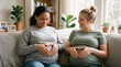 © haris - Two pregnant women of different ethnicities sitting on a couch, smiling and forming heart shapes on their bellies.