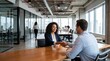 © haris - A smiling businesswoman and a male colleague shaking hands across a wooden table in a bright, modern open-plan office environment with large windows and other employees in the background.