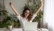 © Selvi - A cheerful woman with brown hair and glasses celebrates with raised arms at her home office desk, wearing a white shirt, with a laptop and green plants in a bright room