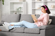 © Pixel-Shot - Pretty young woman with headphones and book listening to audiobook on sofa in living room