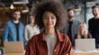 © Muntazim - Confident young woman of mixed ethnicity stands proudly in a modern office with colleagues in the background, showcasing teamwork and collaboration.
