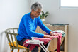 © Gatot - Focused Elderly Asian Man, Wearing Blue Shirt, Smooths Clothes on an Ironing Board Seated in Bright