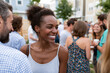 © lashkhidzetim - Joyful woman smiles at outdoor gathering. She wears a light tank top, hair styled in a bun. Surrounding people engage in casual conversation. Background shows trees, buildings, and warm daylight