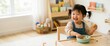 © Xenia - Happy Asian toddler girl eating a messy snack at a small wooden table. Smiling child in denim overalls in a bright playroom. Wide banner with copy space for text