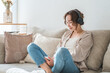 © buritora - A relaxed woman sitting on a sofa at home, listening to music with headphones on.