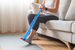 © Nanci - Woman sitting on a sofa performing foot rehabilitation exercises with a blue resistance band, focusing on recovery and strengthening muscles for physical therapy at home