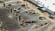 © AmazingAerialAgency - Aerial view of a construction site featuring heavy machinery, excavators, rollers, and various utility trucks parked on a paved lot near a large warehouse building in the United States.