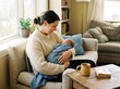 © ari - A mother lovingly cradles her newborn baby wrapped in a blue blanket while sitting on a couch by a window, with a book and mug on a table nearby