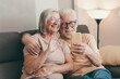 © luciano - Senior Couple Hugging on Sofa During Happy Video Call With Smartphone at Home
