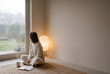 © MystoganCreative - A woman sits cross-legged on a rug, looking out a large window at a garden, with a book and mug nearby, in a cozy room.
