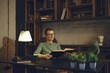 © Aleksei Isachenko/imageBROKER - A person sits comfortably in a modern workspace, immersed in a book. Soft lamp light creates a warm atmosphere. Green plants and rustic shelves add character to the relaxing environment