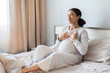 © Anastasiya - Pregnant black woman sitting on bed and practicing calm breathing with hands on her chest, showing prenatal relaxation and wellness at home