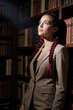 © f_bossa - Woman with red braids looks upward while standing beside wooden bookshelves in a dark library. Soft side lighting highlights her face and textured blazer against the dim interior.