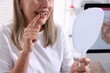 © New Africa - Senior woman looking in mirror at her teeth with dental implant at clinic, closeup