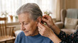 © solom - Caregiver hands helping adjust and fasten a small digital hearing aid behind the ear of an elderly senior woman at home or retirement nursing facility