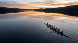 Aerial view of a rowing shell with a crew of eight rowers and a coxswain gliding across a calm, reflective lake during a vibrant sunrise surrounded by dense evergreen forest hills.
