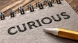 © NingPhattraphorn - A close-up of a spiral notebook with the word 'CURIOUS' written on the page and a sharpened yellow pencil lying nearby, artistic work text banner