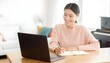 © Wankatsu - Cheerful young Asian woman wearing a pink sweater and earphones taking notes while looking at a laptop in a bright home office modern minimalist lifestyle concept