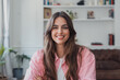 © Daniel - Cheerful young woman with long brown hair smiling confidently while looking at the camera, posing inside a bright, modern home interior