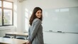 © Dyeus - Smiling young professional woman standing in a modern classroom, looking back cheerfully. Happy teacher or educator in an educational setting.