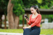 © Tj - Young Asian woman is sitting outdoors in a park, holding a smartphone and a coffee cup, focusing on her device for communication, social media, or remote work activities