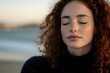© Yuliia - Young woman with curly reddish-brown hair and fair complexion wearing dark sweater, eyes closed in peaceful expression by ocean at sunset, contemplative mood for wellness and lifestyle content.