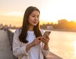 © Selalu - Young woman in casual attire using smartphone on walkway by serene water during sunset