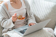 © buritora - A woman sits on a sofa, sipping tea or herbal tea while looking at her computer.