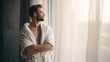 © Yaryna - Man in bathrobe looking out window, contemplating future, morning light. A thoughtful man wearing a white terry cloth robe stands by a window, bathed in soft morning light, arms crossed