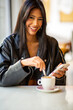 © Paula - Smiling woman using smartphone and drinking coffee in cafe