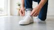 © Celt Studio - Casual young man wearing blue jeans kneeling on grey concrete floor tying shoelaces on stylish crisp white leather sneakers before going outside